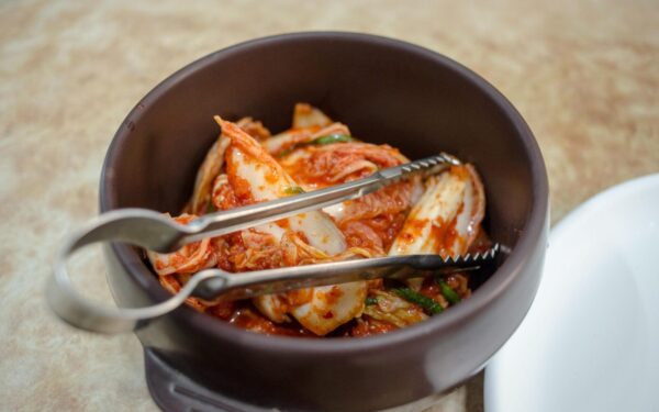 Close-up of traditional Korean kimchi in a dark ceramic bowl with tongs.