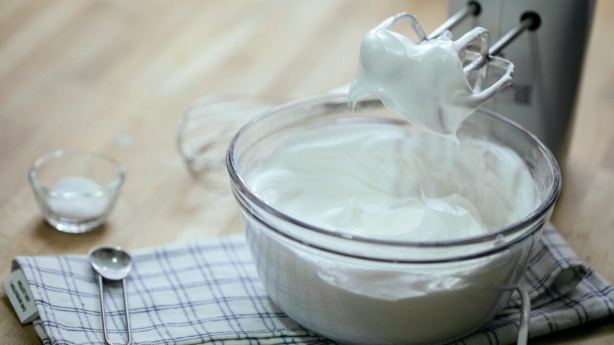 Arrangement of tasty fresh whipped cream in glass bowl near mixer spoon on towel on kitchen table