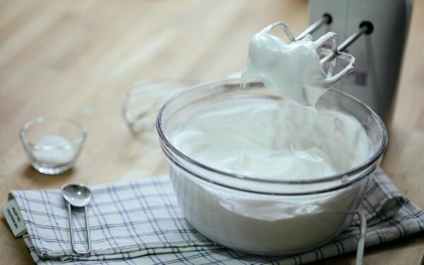 Arrangement of tasty fresh whipped cream in glass bowl near mixer spoon on towel on kitchen table