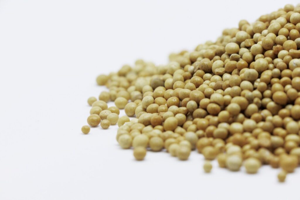 A close-up image showing a pile of brown mustard seeds on a white surface, highlighting their texture and color.